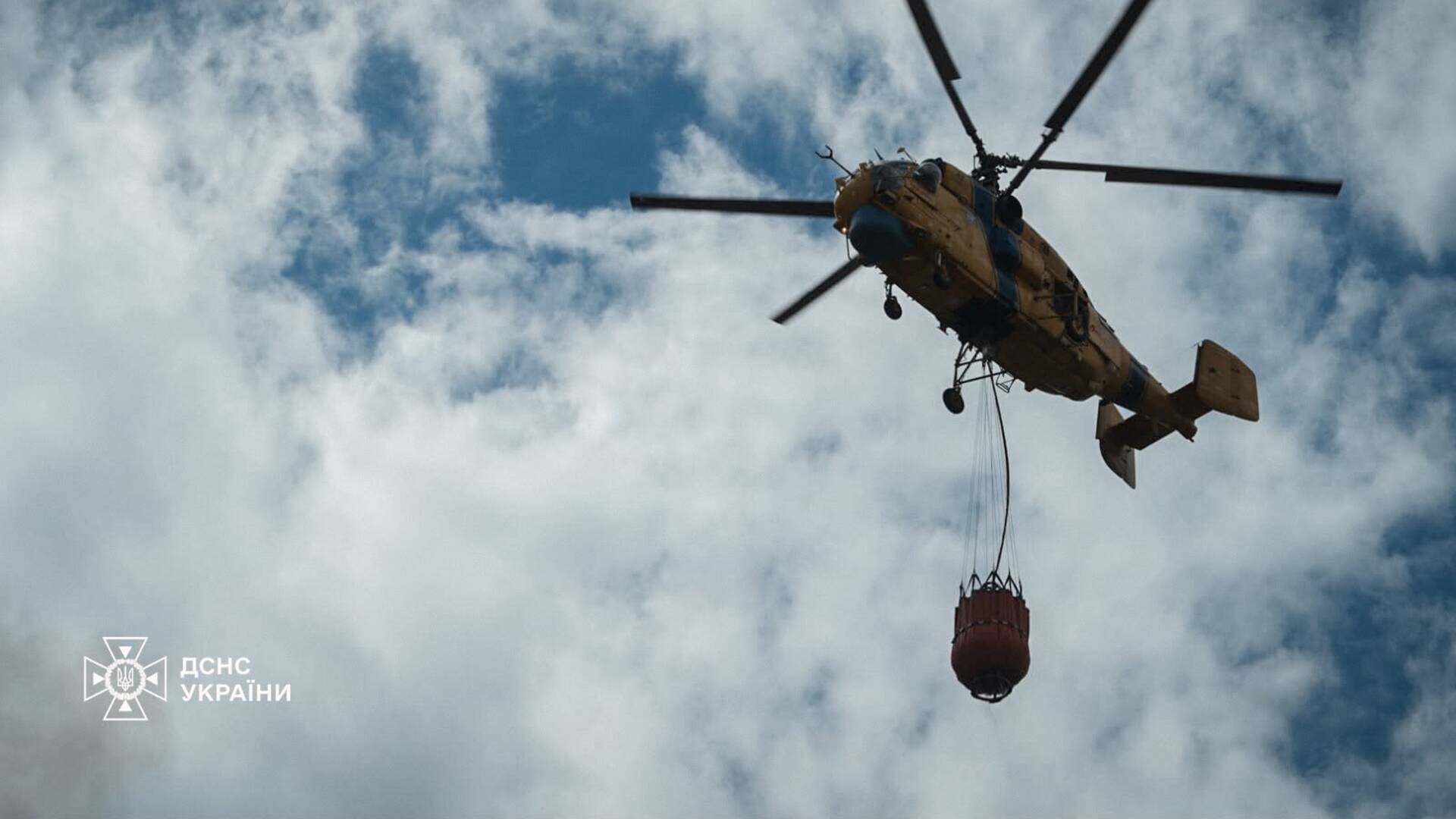 Вертоліт авіації ДСНС України з водяним відром під фюзеляжем летить у хмарному небі до осередку пожежі