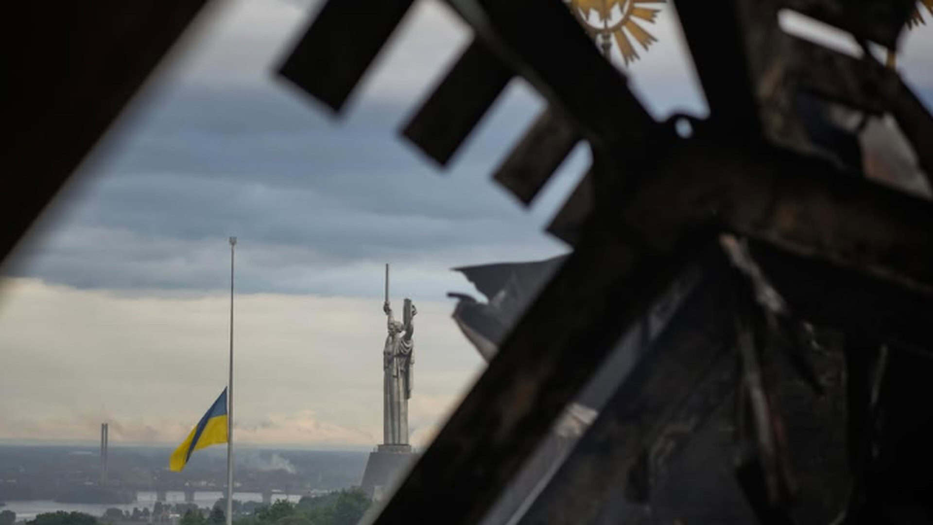 Монумент Батьківщина-Мати і приспущений прапор України крізь обгорілу конструкцію будівлі після обстрілу
