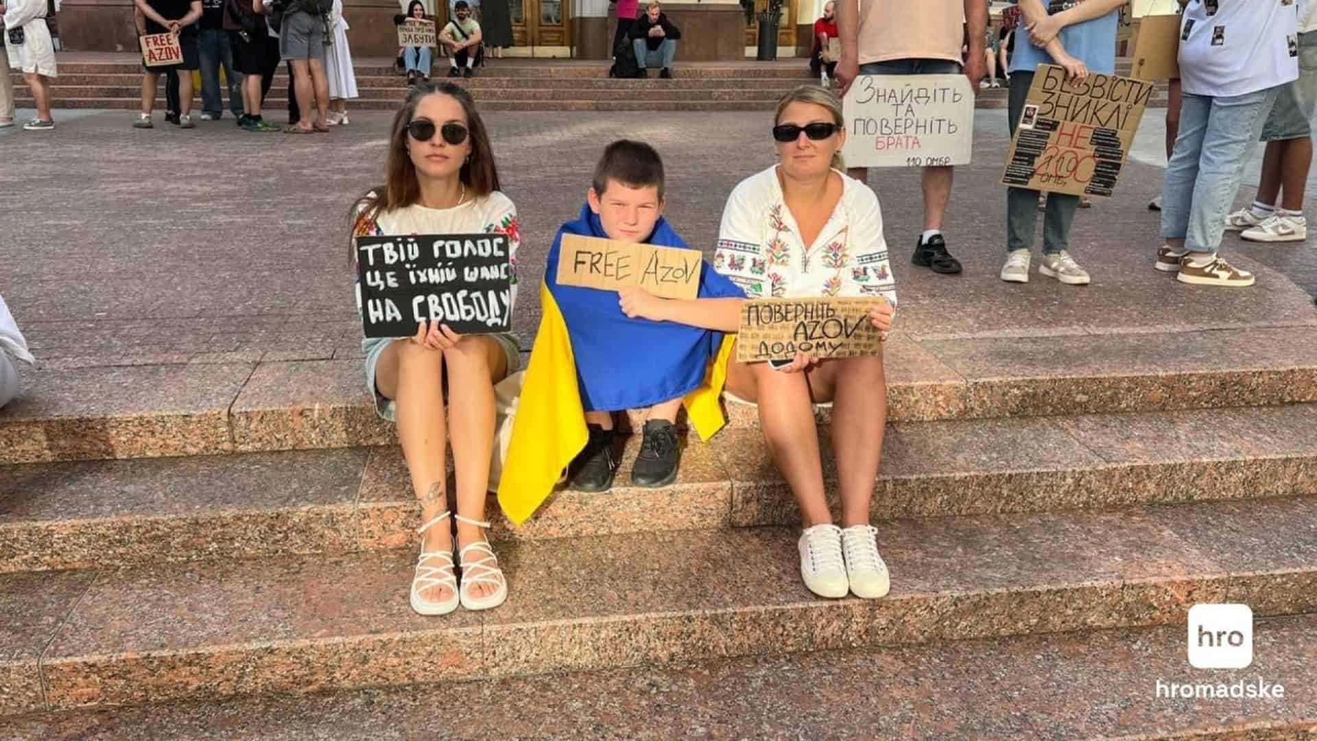 Жінка, хлопчик у синьо-жовтому прапорі та жінка тримають плакати «Free Azov» і «Поверніть Azov додому» на сходах будівлі в Києві