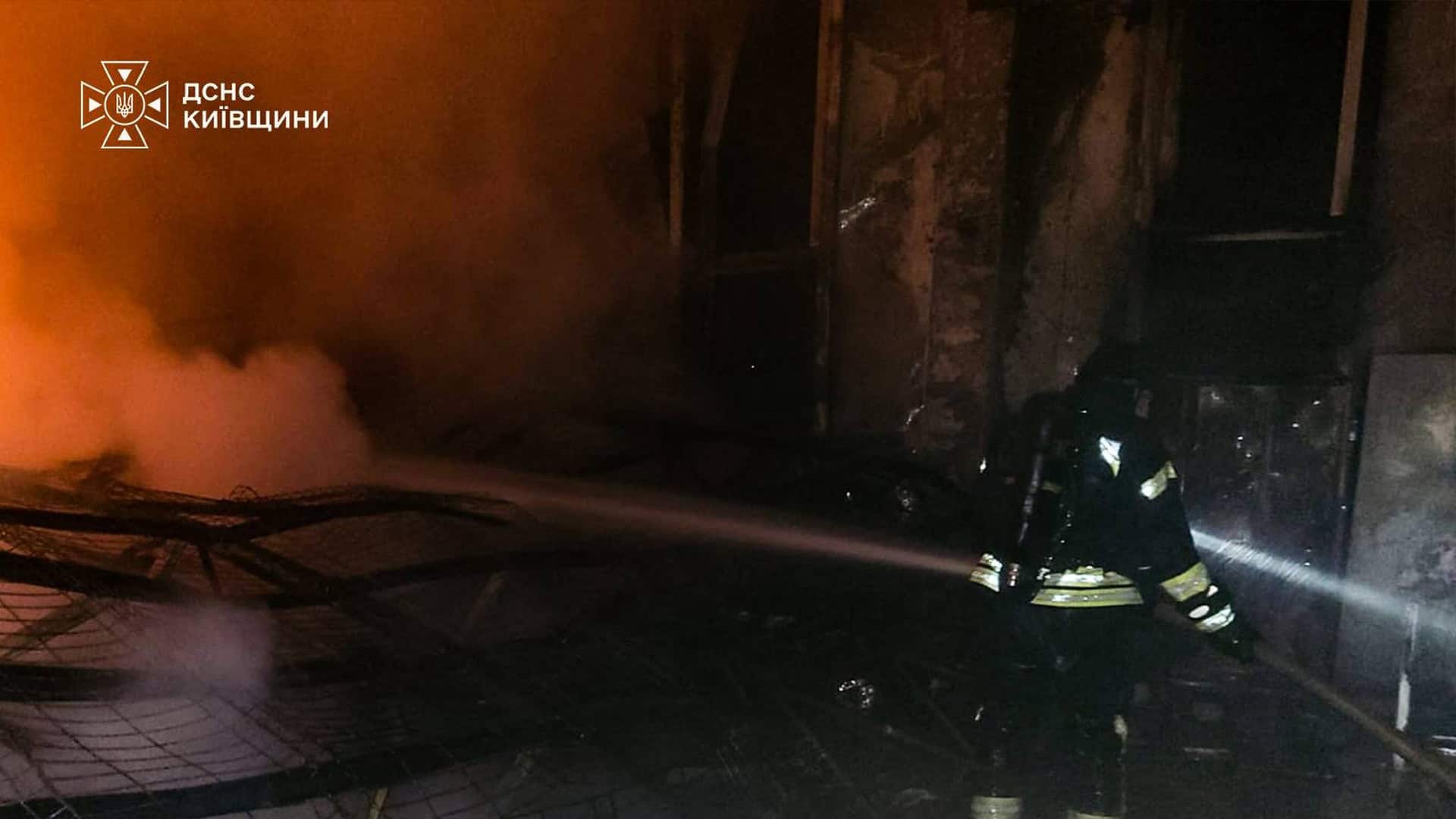 Рятувальник ДСНС гасить пожежу у пошкодженій будівлі на Київщині, навколо густий дим і уламки конструкцій після нічної атаки.