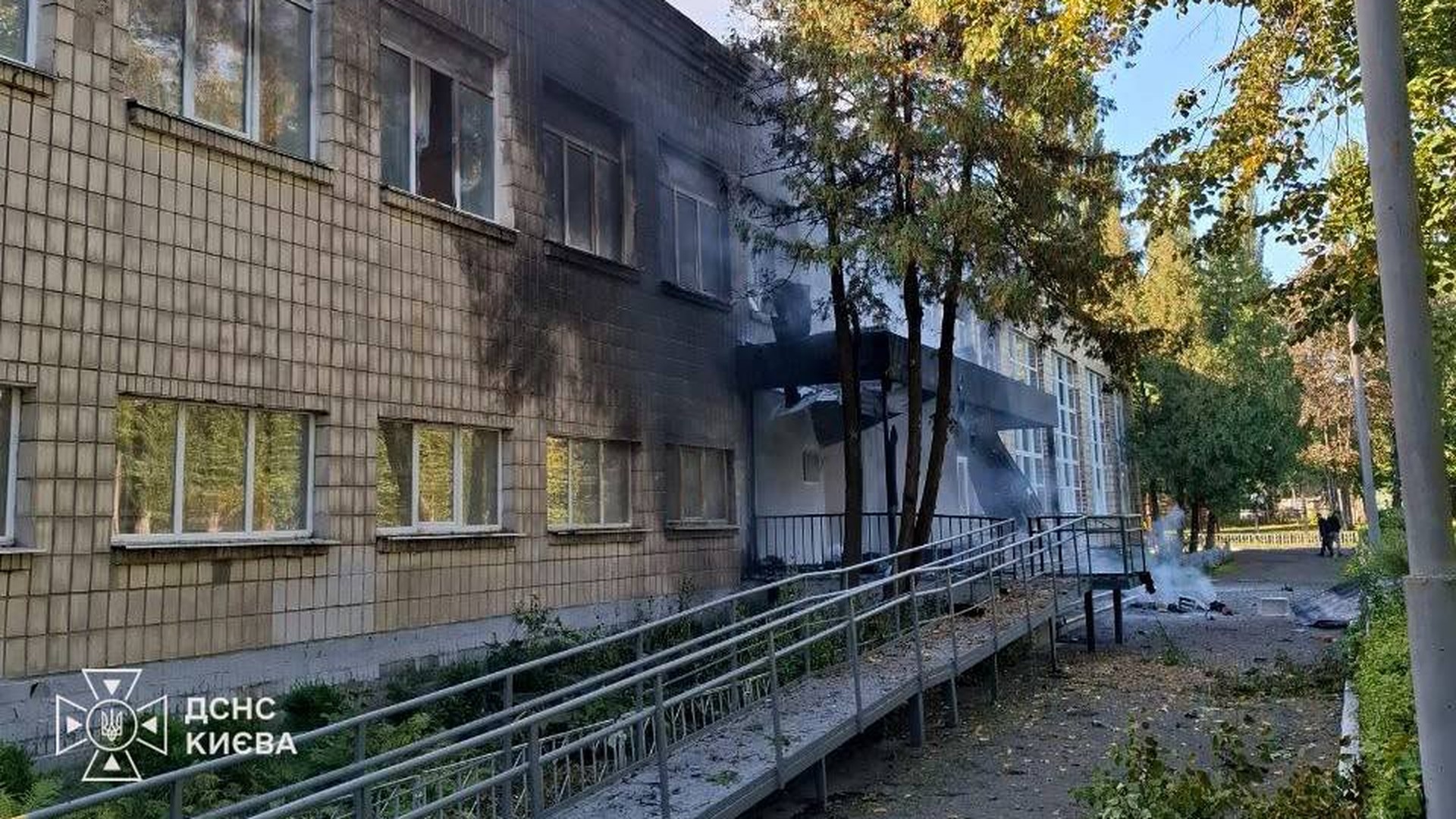 Закопчений фасад закладу освіти в Києві з вибитими вікнами, біля входу ще дим, пандус засипаний уламками