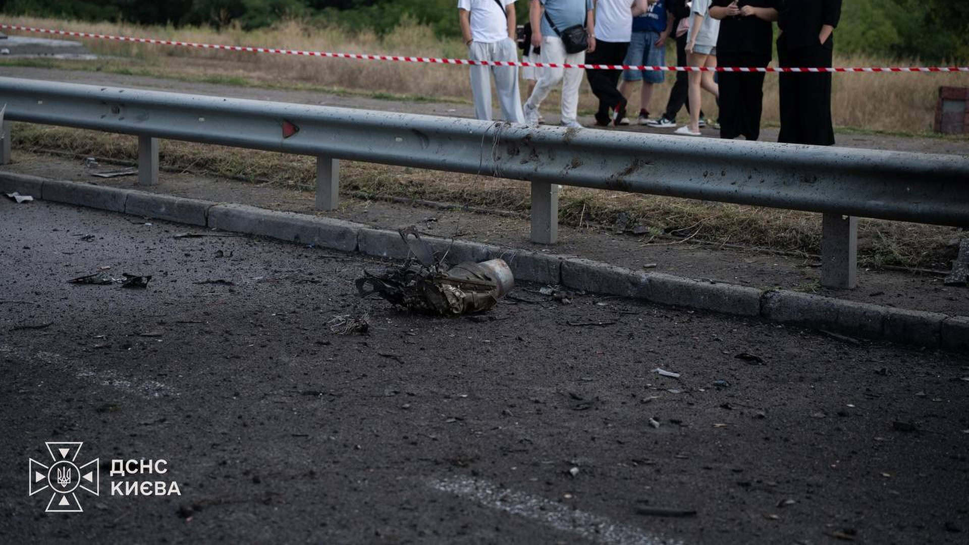 Обгорілий металевий уламок дрона лежить на асфальті біля дорожнього відбійника, за огороджувальною стрічкою стоять люди