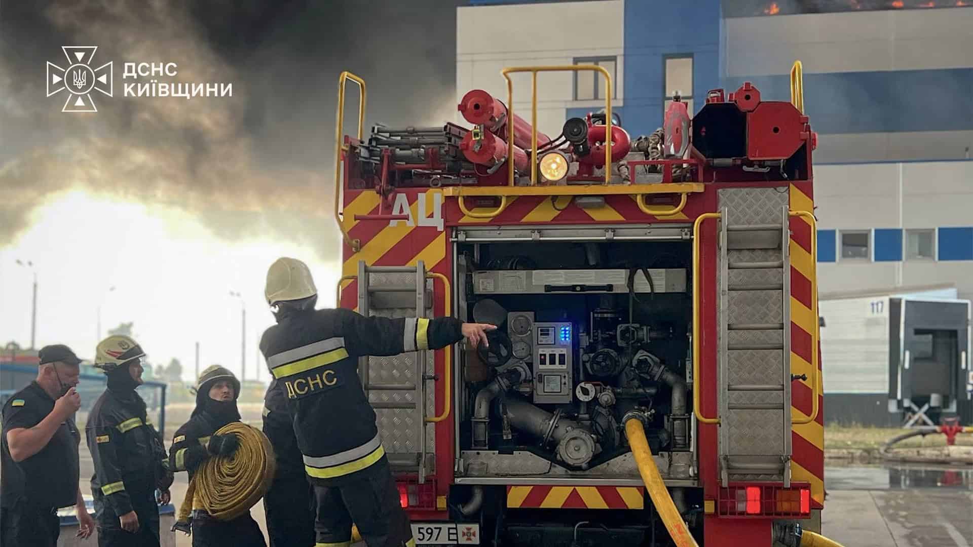 Рятувальники ДСНС розмотують пожежний рукав біля автоцистерни на тлі великого стовпа чорного диму й полум'я над будівлею