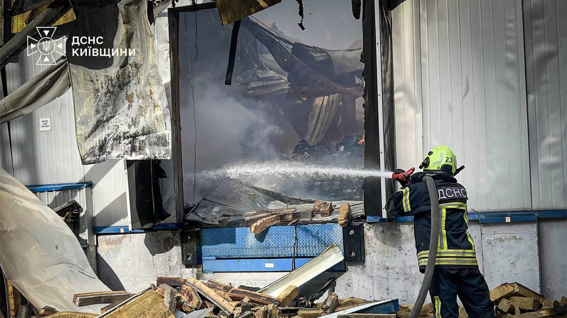 Рятувальник ДСНС подає струмінь води в отвір зруйнованої стіни складу, навколо розкидані уламки конструкцій і дерев'яні палети