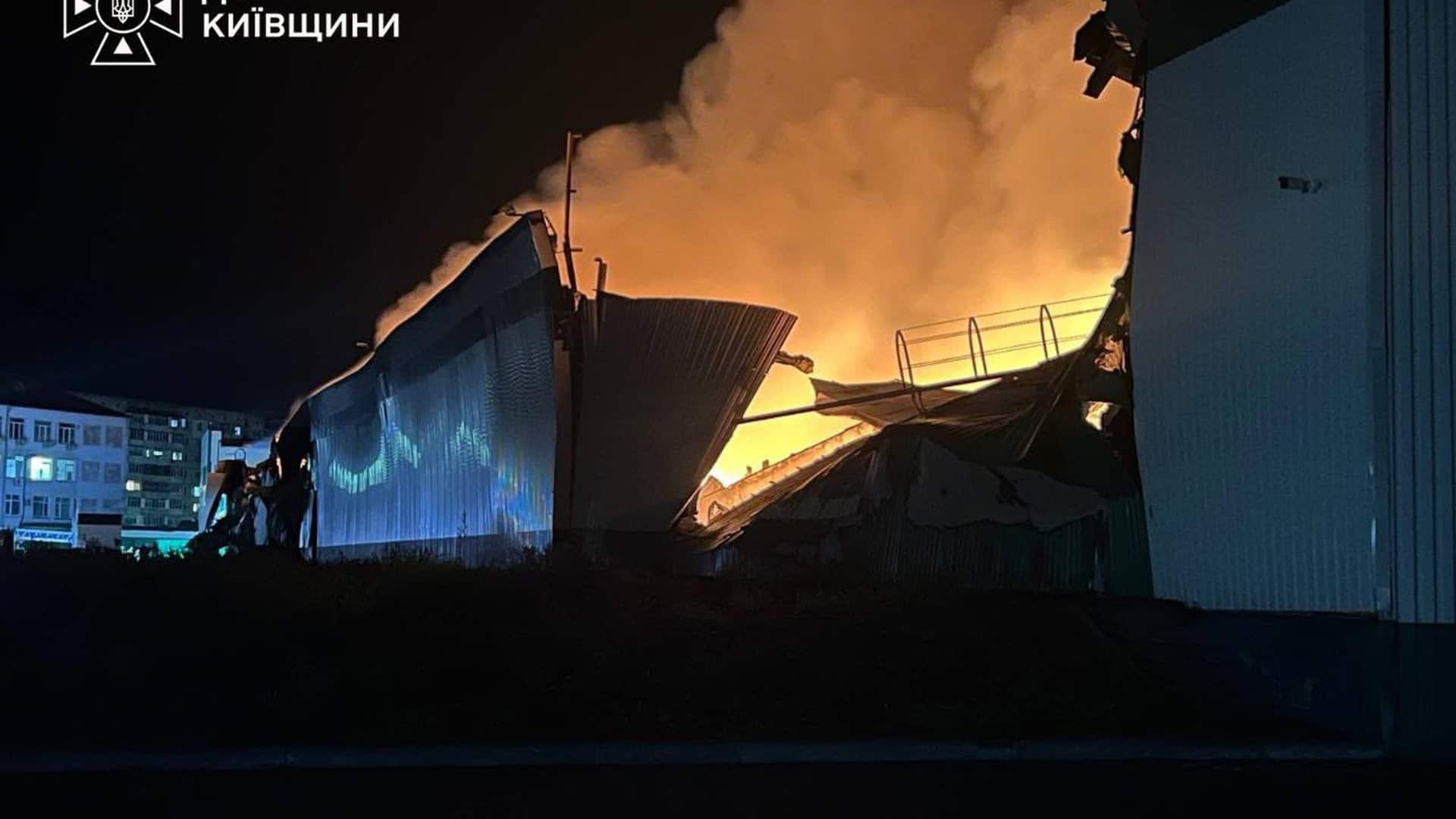 Зруйнована й охоплена вогнем покрівля складського ангару вночі, потужне полум'я освітлює навколишні будівлі