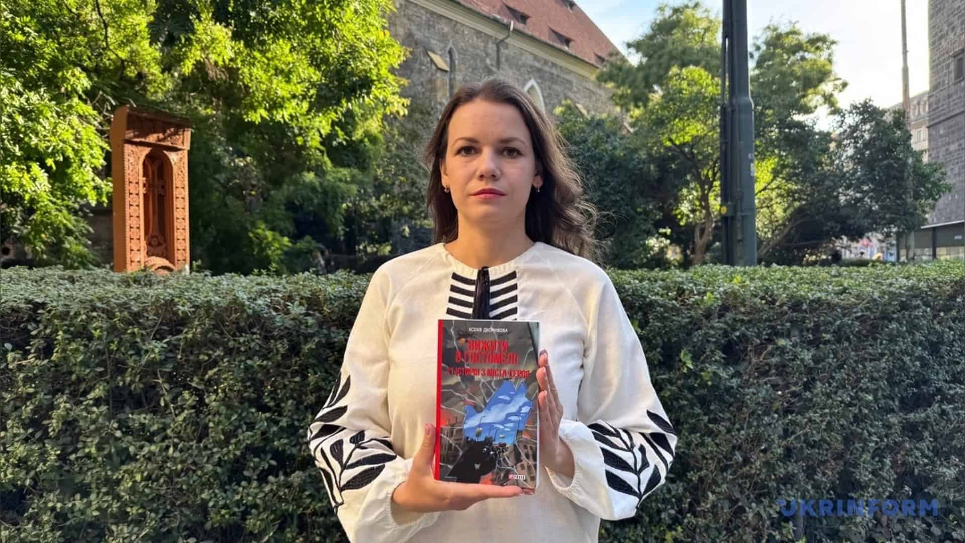Ксенія Дворнікова у вишиванці тримає книгу «Вижити в Гостомелі» на тлі живоплоту в Празі