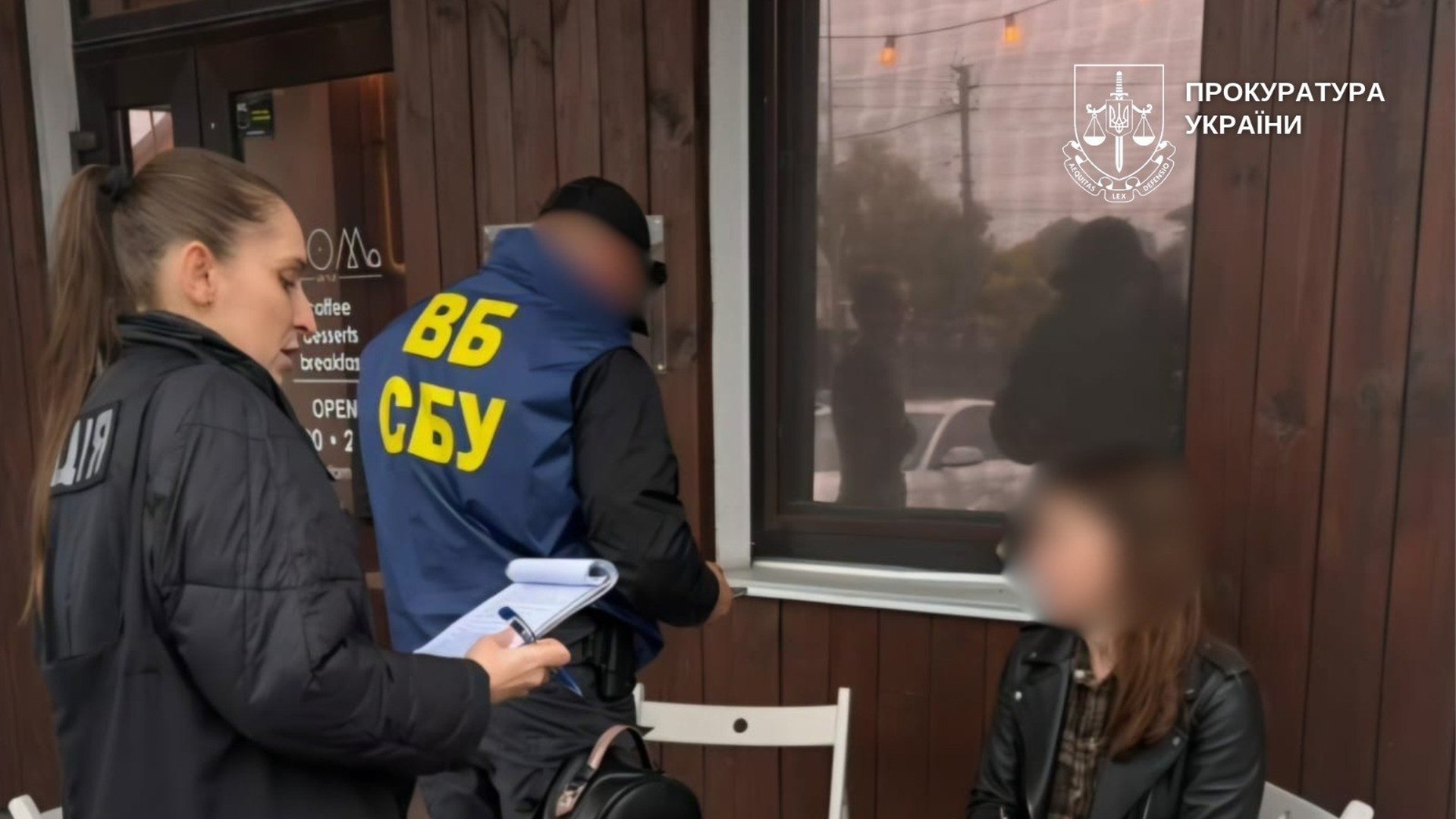 Поліцейська з блокнотом і співробітник СБУ біля жінки з розмитим обличчям на терасі кав’ярні під час затримання в Києві