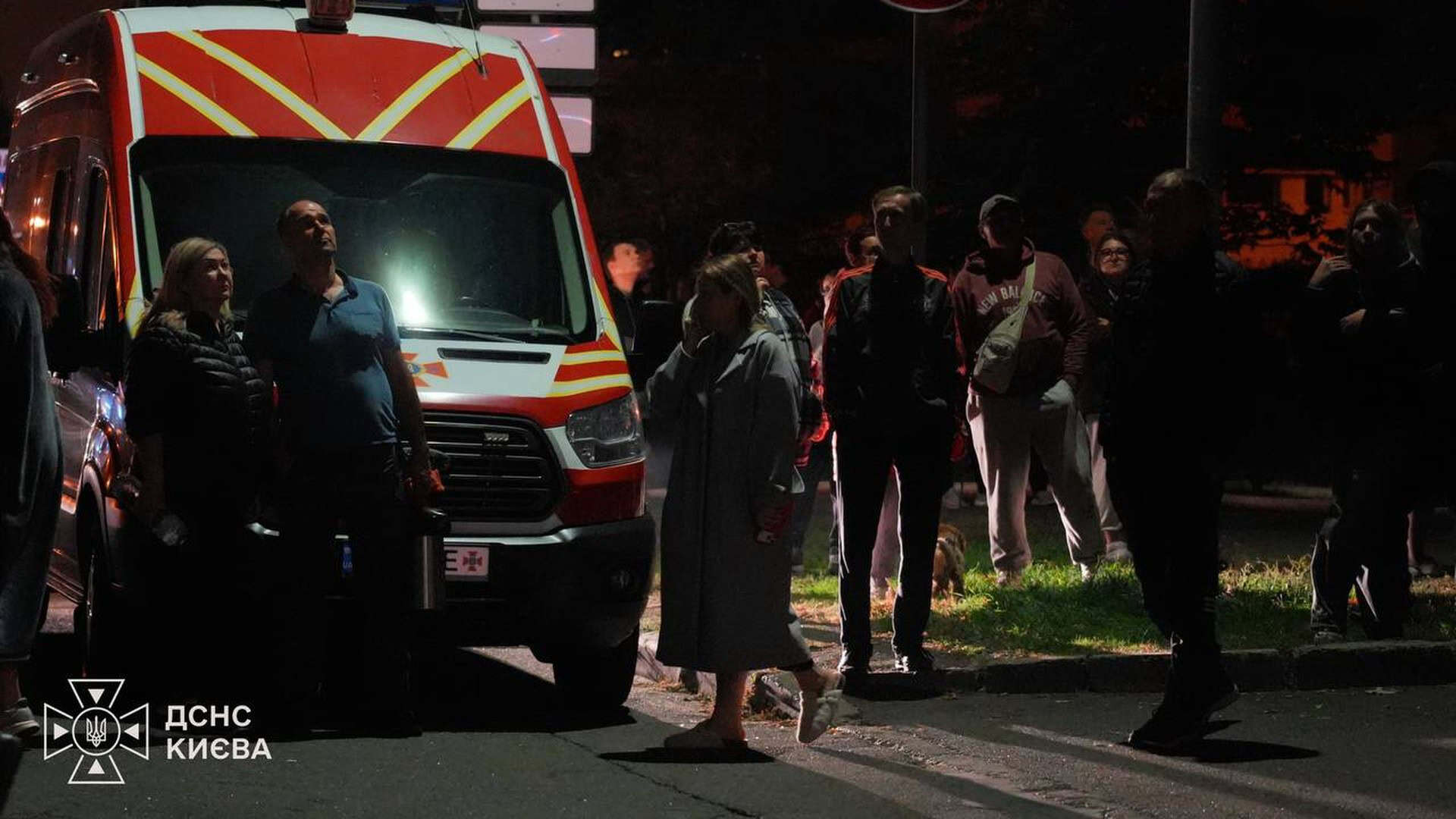 Мешканці Києва вночі стоять біля автомобіля ДСНС на вулиці під час ліквідації наслідків російської атаки