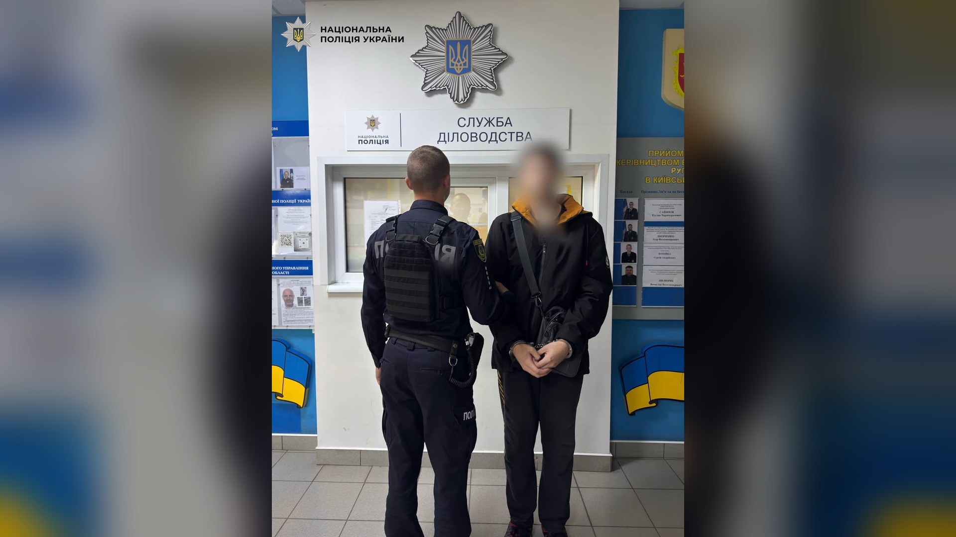 Поліцейський веде затриманого в кайданках із заблюреним обличчям у відділі поліції на Київщині
