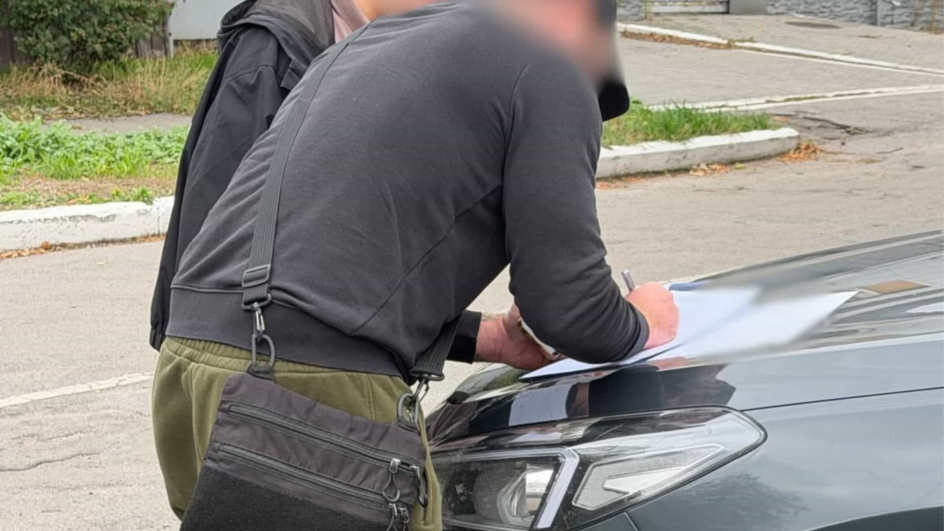 Чоловік із заблюреним обличчям підписує документи на капоті автомобіля у справі про невстановлений модульний будинок у Києві