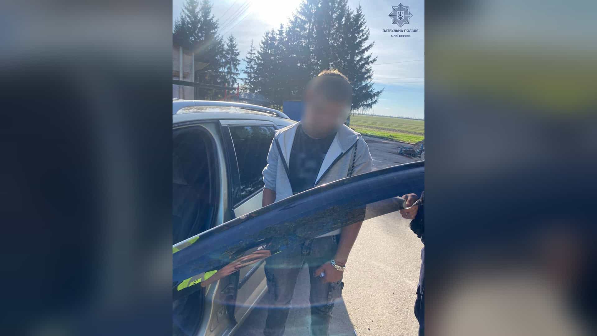 Чоловік із розмитим обличчям стоїть біля автомобіля під час перевірки документів, праворуч видно руку патрульного поліцейського