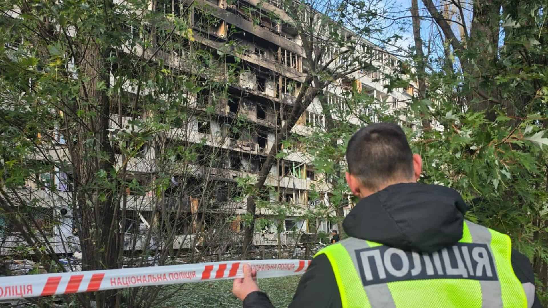 Поліцейський тримає стрічку огородження біля обгорілої багатоповерхівки в Деснянському р-ні Києва після російського удару