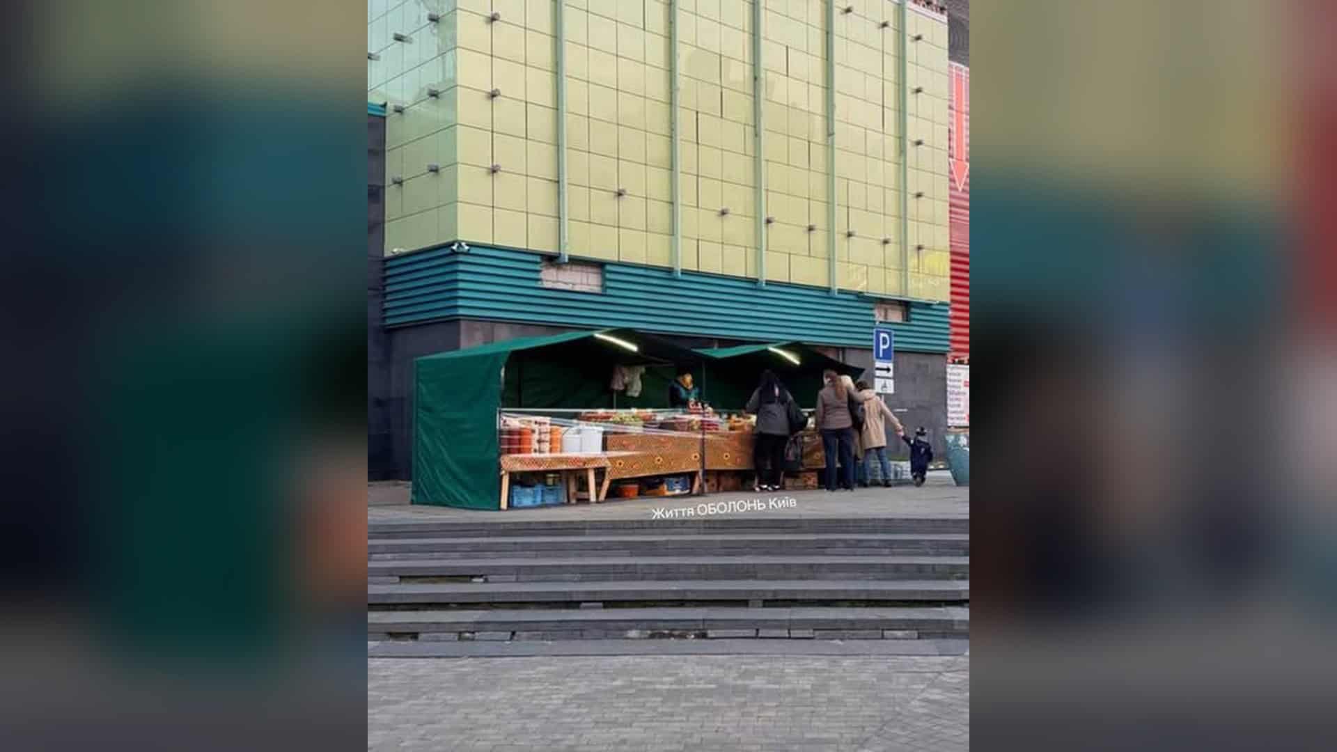 Зелений торговельний намет із прилавками біля торговельної будівлі в Оболонському р-ні, поряд стоять покупці