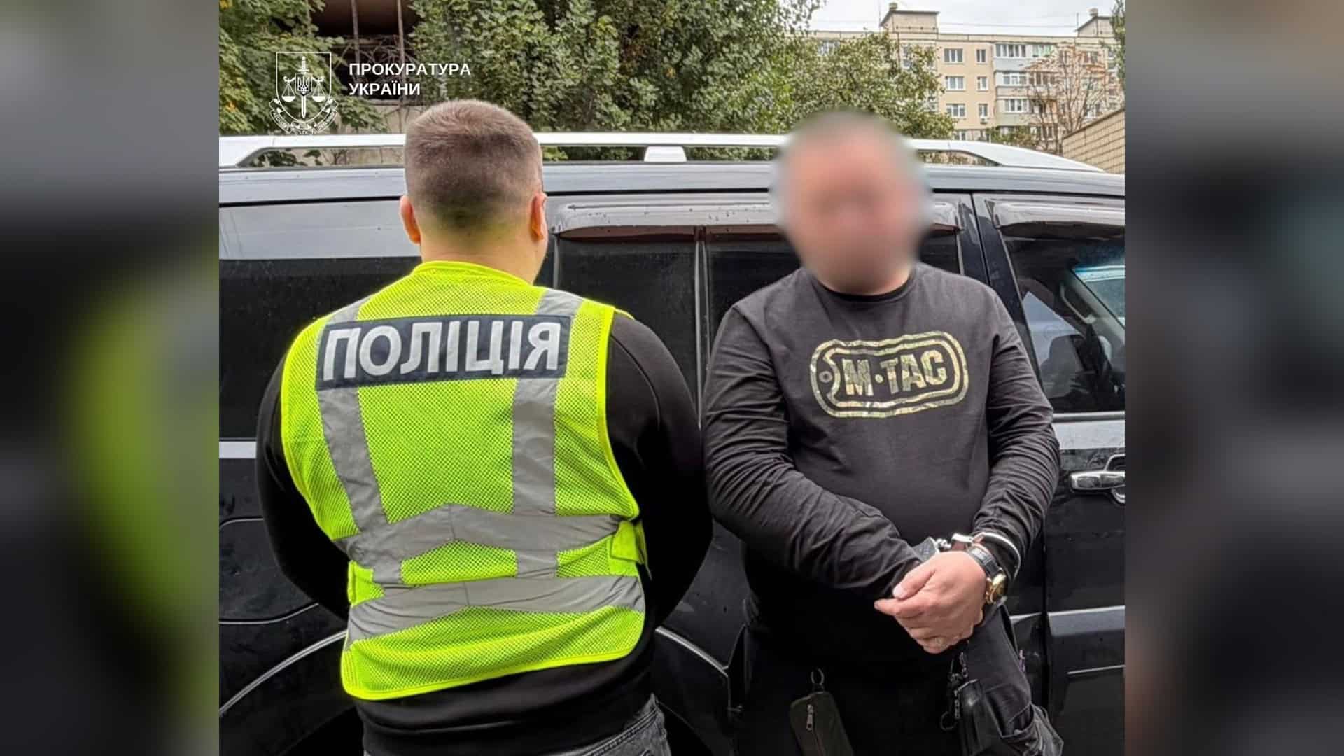 Поліцейський у жилеті поруч із затриманим чоловіком у кайданках із розмитим обличчям біля авто в Києві