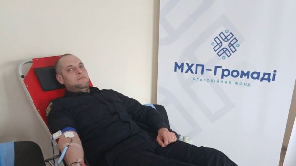 Поліцейський у формі здає кров під час донорської акції на підтримку поранених військових, банер благодійного фонду МХП-Громаді на фоні