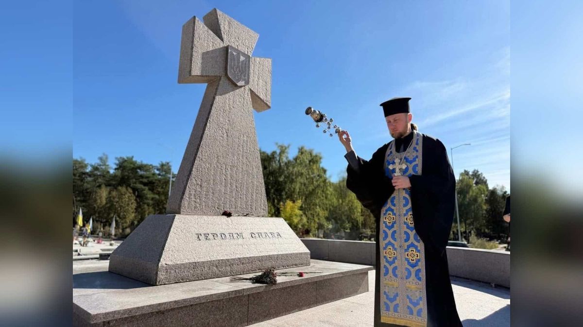 Священник із кадилом і хрестом біля кам’яного козацького хреста з написом «Героям слава» на кладовищі, квіти на постаменті
