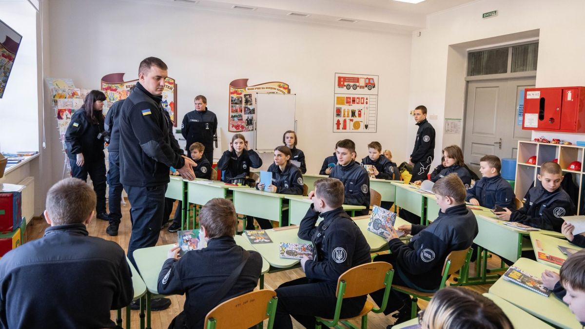 Діти у формі Київської дитячої залізниці сидять за партами в Класі безпеки ДСНС, рятувальник проводить інтерактивний урок з правил безпеки