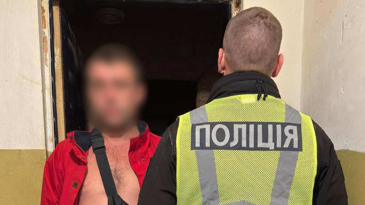 Поліцейський у жилеті з написом «Поліція» поруч із затриманим чоловіком у червоній куртці біля дверей квартири в Києві