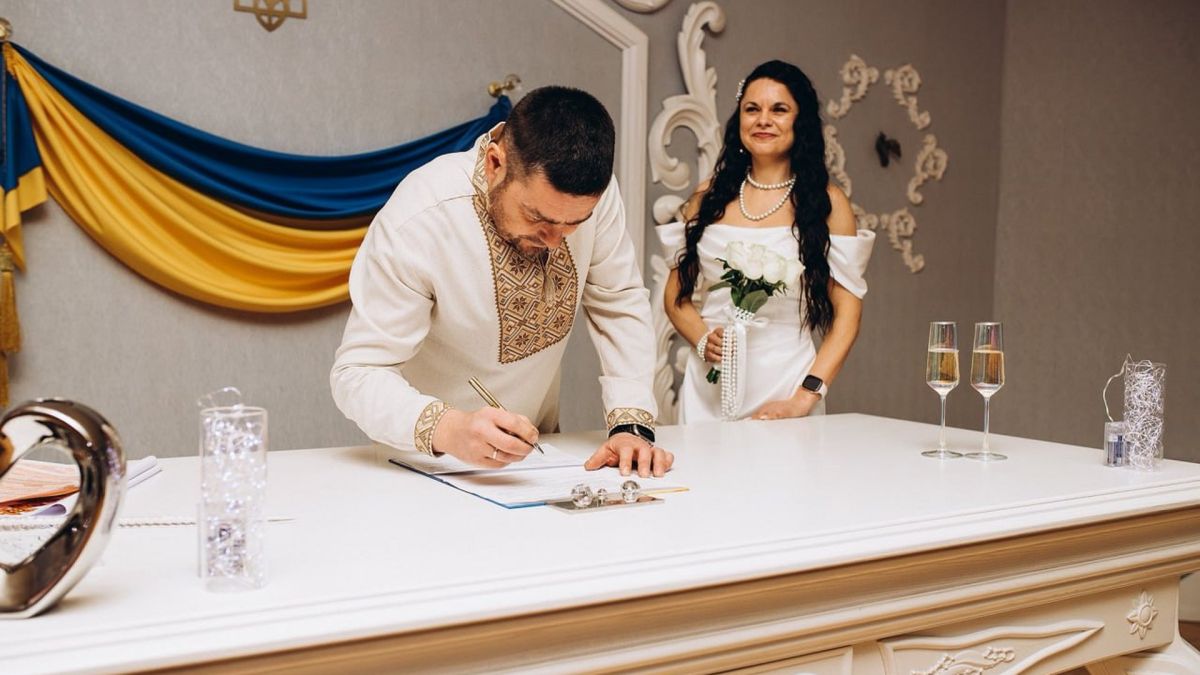 Наречений у вишиванці підписує документи про шлюб, наречена у білій сукні стоїть поруч із букетом