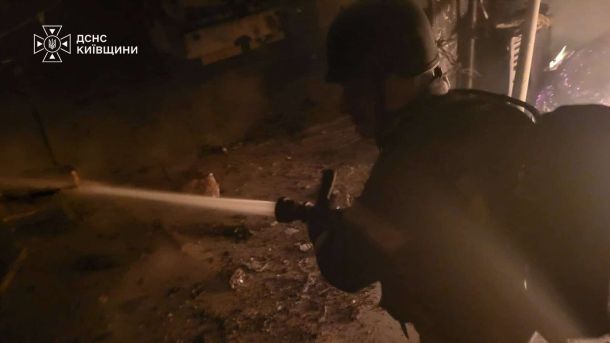 Рятувальник ДСНС у захисному шоломі спрямовує струмінь води на осередок пожежі серед задимлених руїн після нічної атаки на Київщині.
