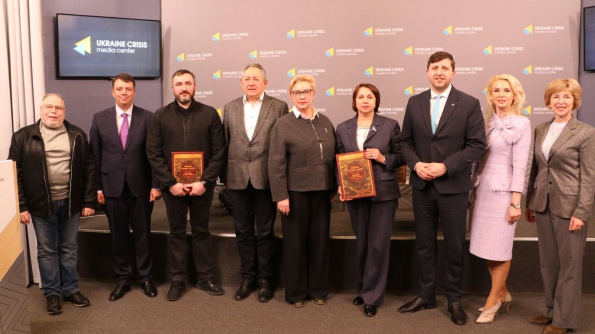 Учасники презентації атласу Київщини тримають два примірники видання в червоних обкладинках на сцені Українського кризового медіа-центру