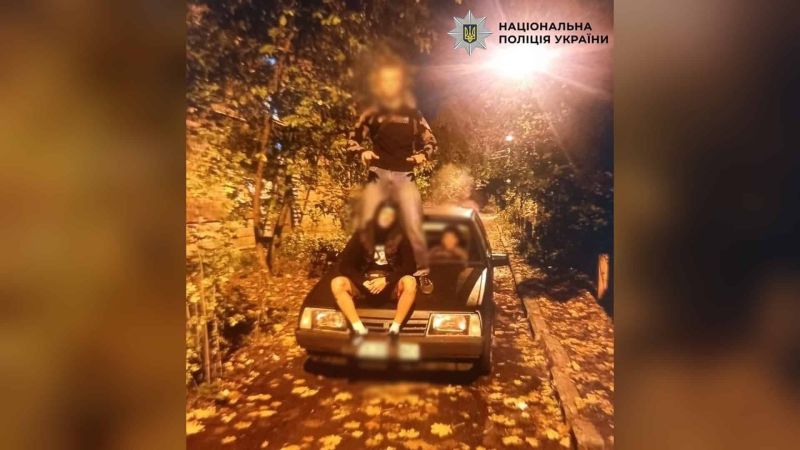 Підлітки позують на капоті та над автомобілем ВАЗ уночі на засипаній листям вулиці, обличчя розмито, логотип Національної поліції України