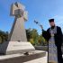 Священник із кадилом і хрестом біля кам’яного козацького хреста з написом «Героям слава» на кладовищі, квіти на постаменті