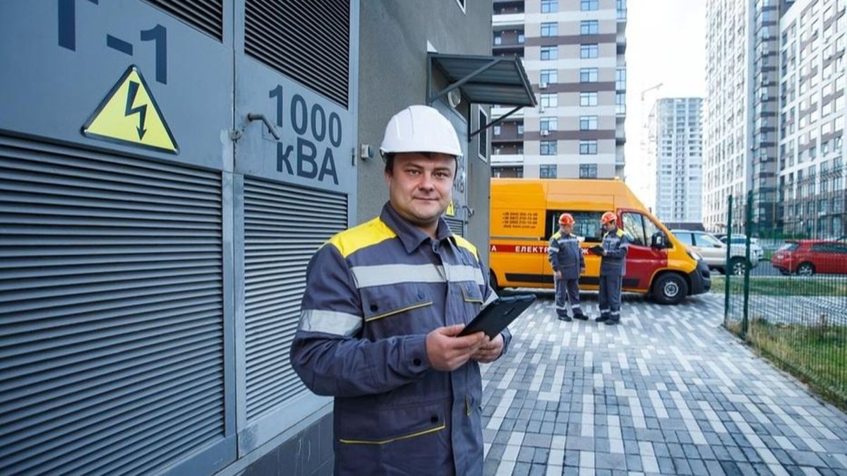Електрик у захисному одязі стоїть біля трансформаторної підстанції, тримаючи планшет