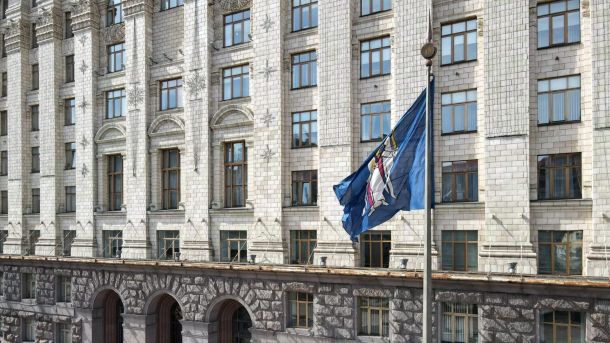 Фасад будівлі КМДА з білої плитки з великими вікнами, перед якою на флагштоці майорить синій прапор із гербом Києва