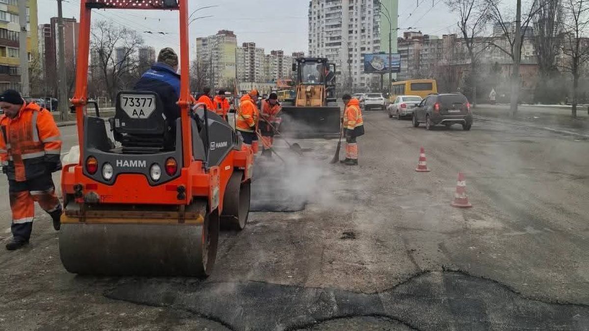 Дорожники у помаранчевих жилетах та каток HAMM проводять ямковий ремонт на вулиці Деснянського району Києва, з-під асфальту йде пара