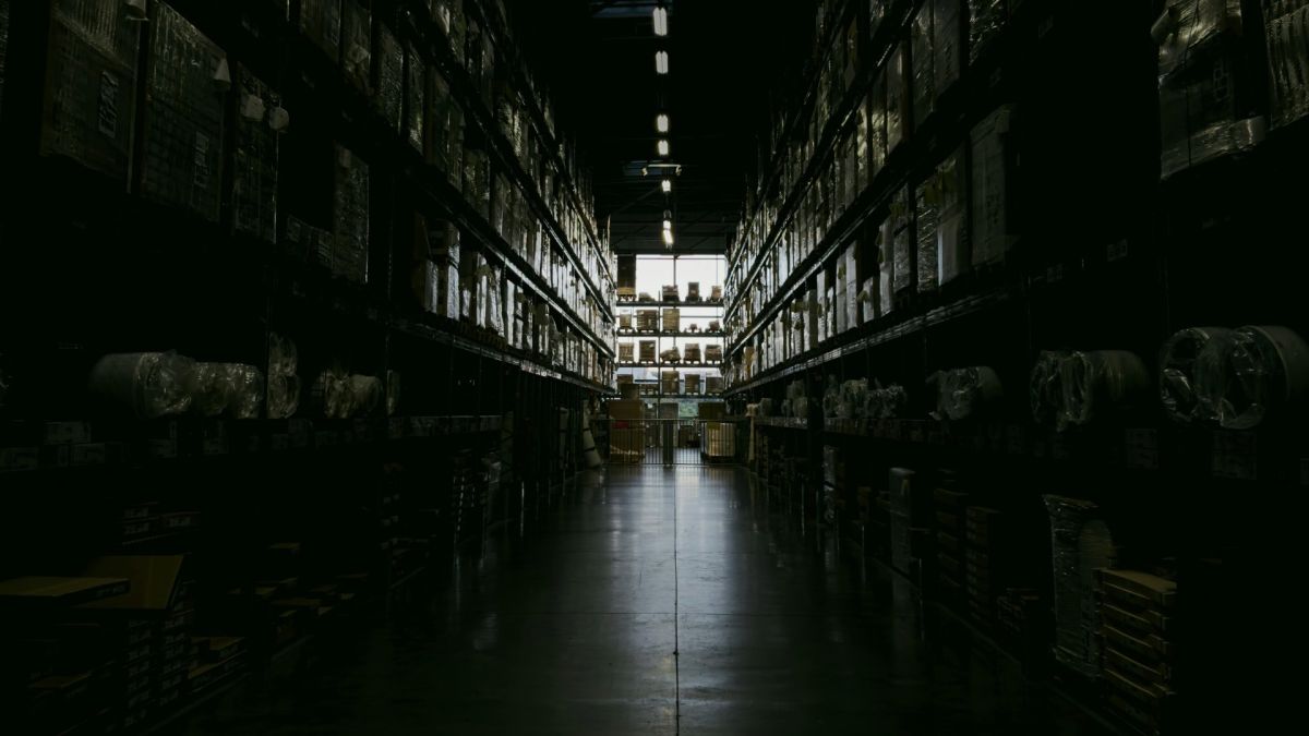 Темний складський прохід із високими стелажами, заповненими коробками та запчастинами, з освітленням у глибині приміщення
