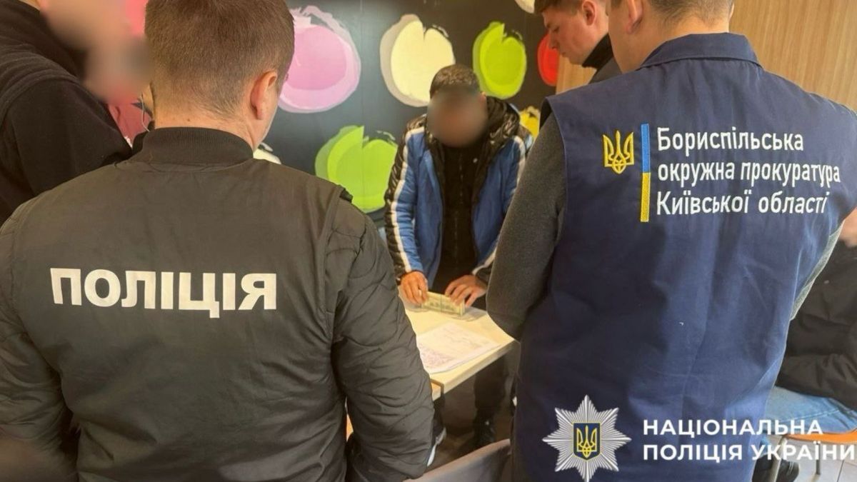 Правоохоронці затримують чоловіка у синій курточці, той стоїть біля стола в тримає папери