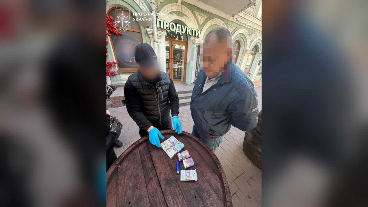 Правоохоронець у рукавичках показує пачки гривень на бочці-столі поруч із підозрюваним, обличчя якого розмите