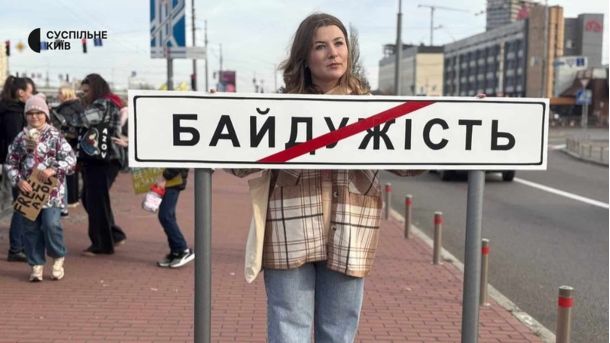 Жінка тримає дорожній знак з написом "БАЙДУЖІСТЬ", перекреслений червоною смугою