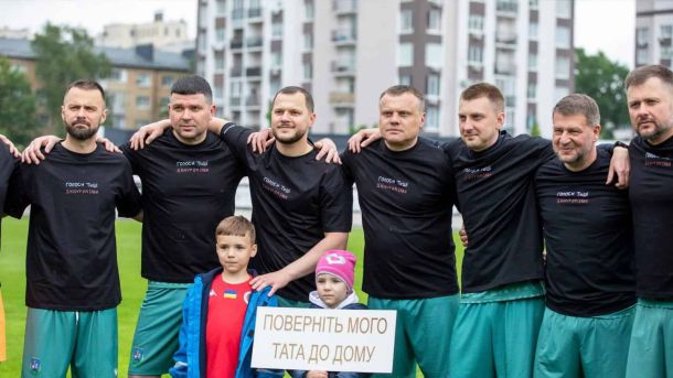 Учасники благодійного матчу «Матч очікування: Кожен голос рятує» в Бучі з дітьми, що тримають плакат «Поверніть мого тата додому»