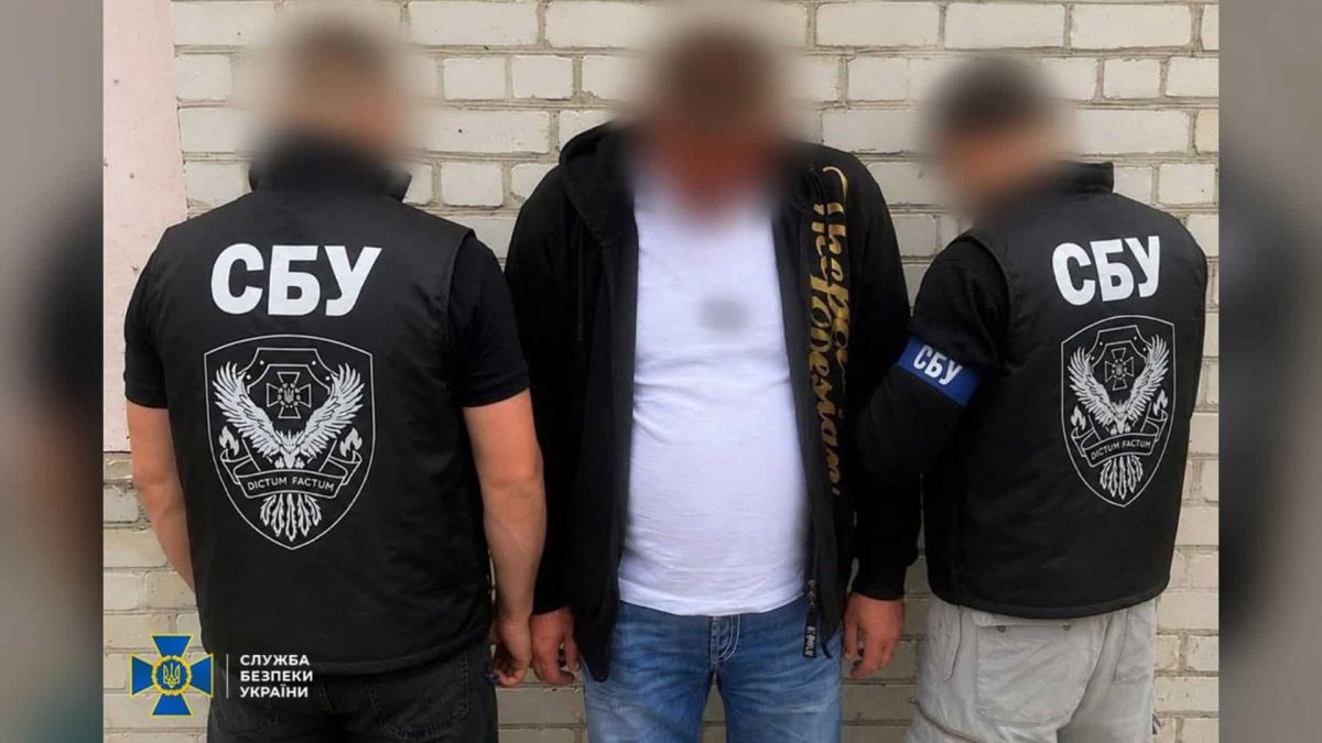 Двоє співробітників СБУ у чорних жилетах біля затриманого чоловіка в білій футболці на тлі цегляної стіни