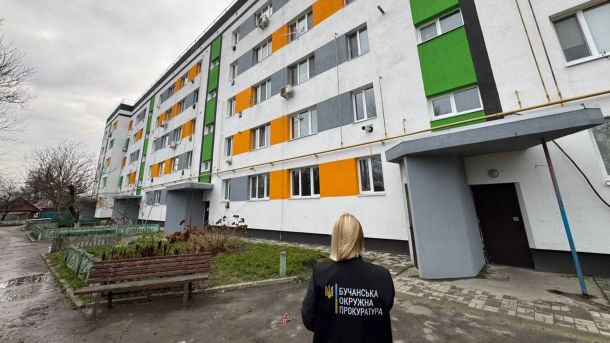 Жінка в чорній куртці із надписом Прокуратура області стоїть біля багатоповерхового житлового будинку