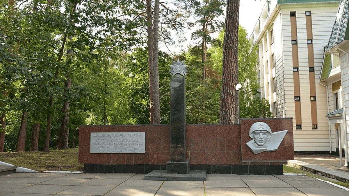 Меморіал з пам'ятною стелою та скульптурою солдата у парку Києва, присвячений історичним подіям та війні, оточений зеленими деревами та сучасною архітектурою.
