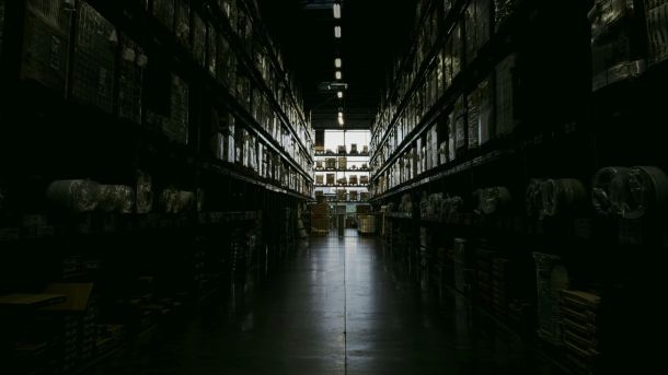 Темний складський прохід із високими стелажами, заповненими коробками та запчастинами, з освітленням у глибині приміщення