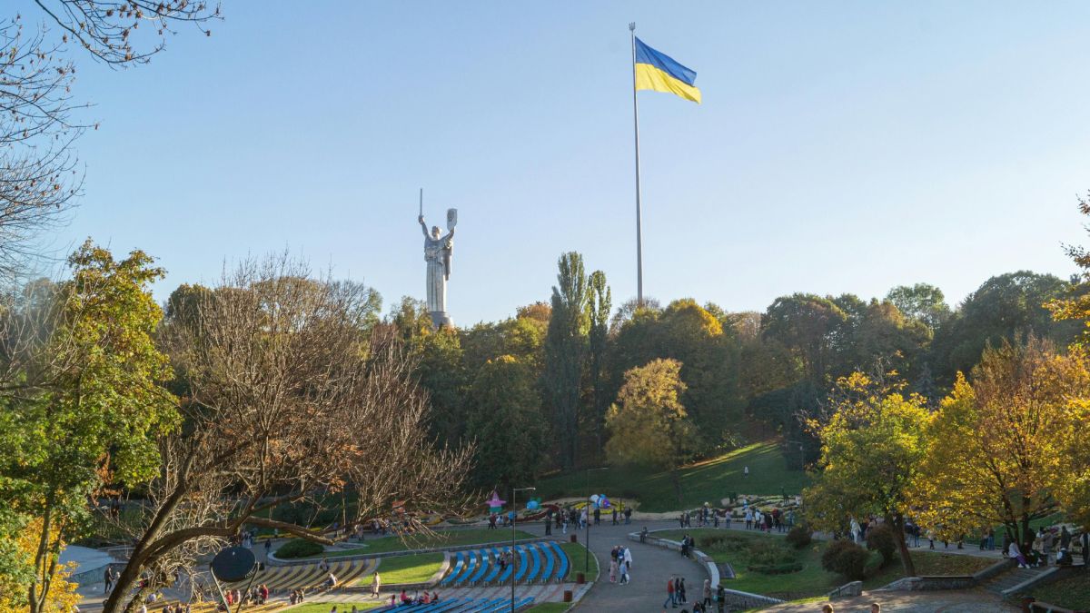 Осінній парк із прогулянковими алеями, монумент Батьківщина-Мати та великий прапор України на тлі блакитного неба в Києві