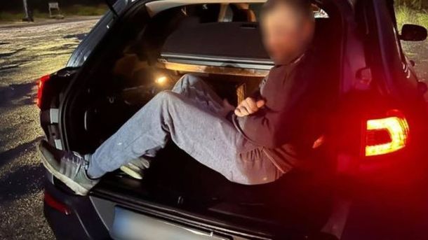 Чоловік у спортивному одязі сидить у багажнику автомобіля з відкритими дверцятами, освітлений червоним світлом задніх фар