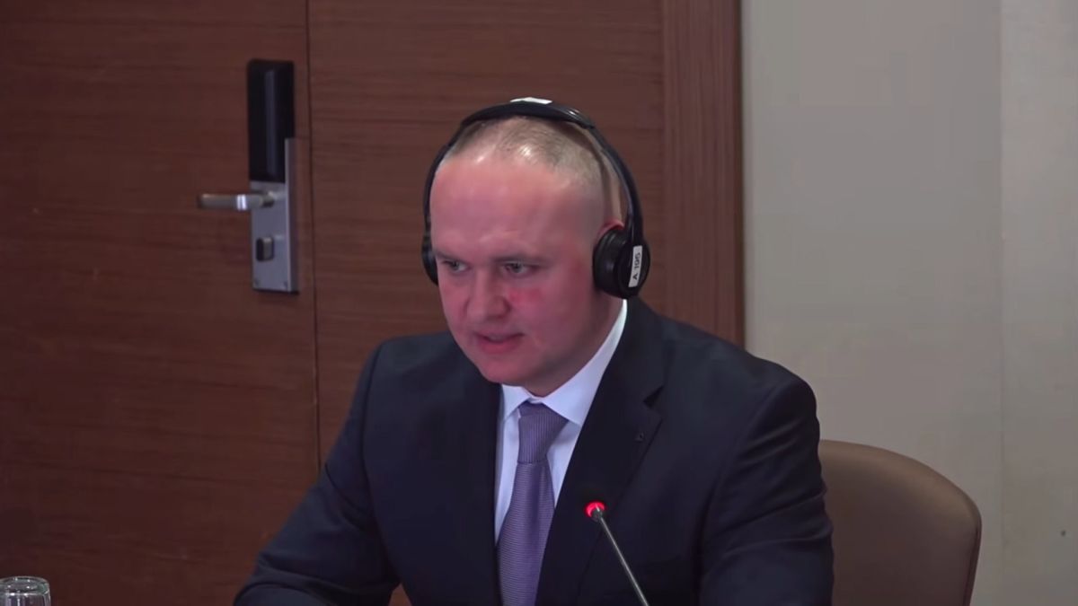 Орест Мандзій на засіданні в навушниках для перекладу виступає перед мікрофоном у діловому костюмі