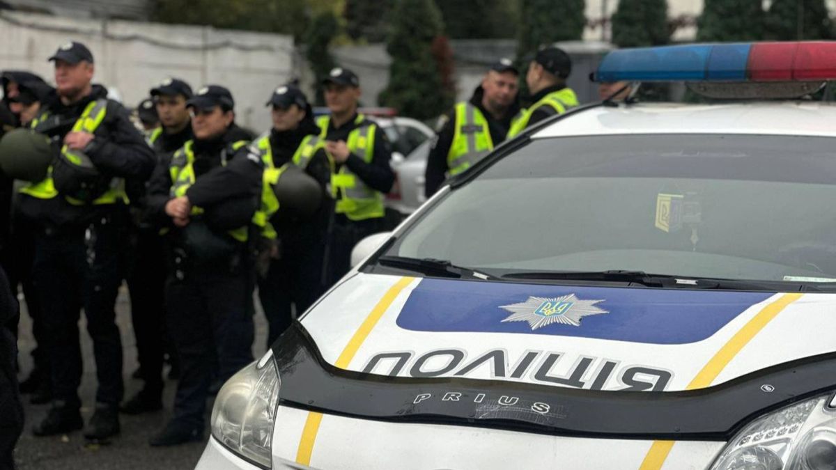 У столиці посилили безпеку: поліція Києва збільшила кількість нарядів через блекаут На передньому плані поліцейський автомобіль із увімкненим проблисковим маячком, на задньому — група поліцейських