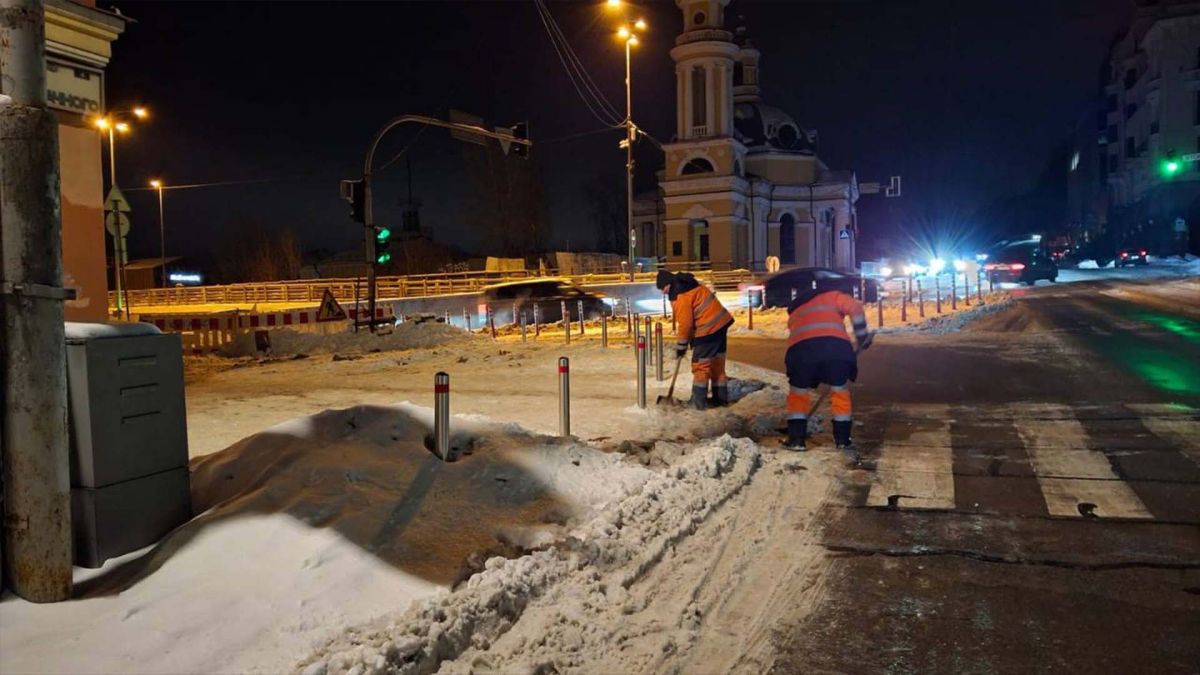 Двоє працівників «Київавтодору» в помаранчових жилетах прибирають сніг та лід біля пішохідного переходу вночі