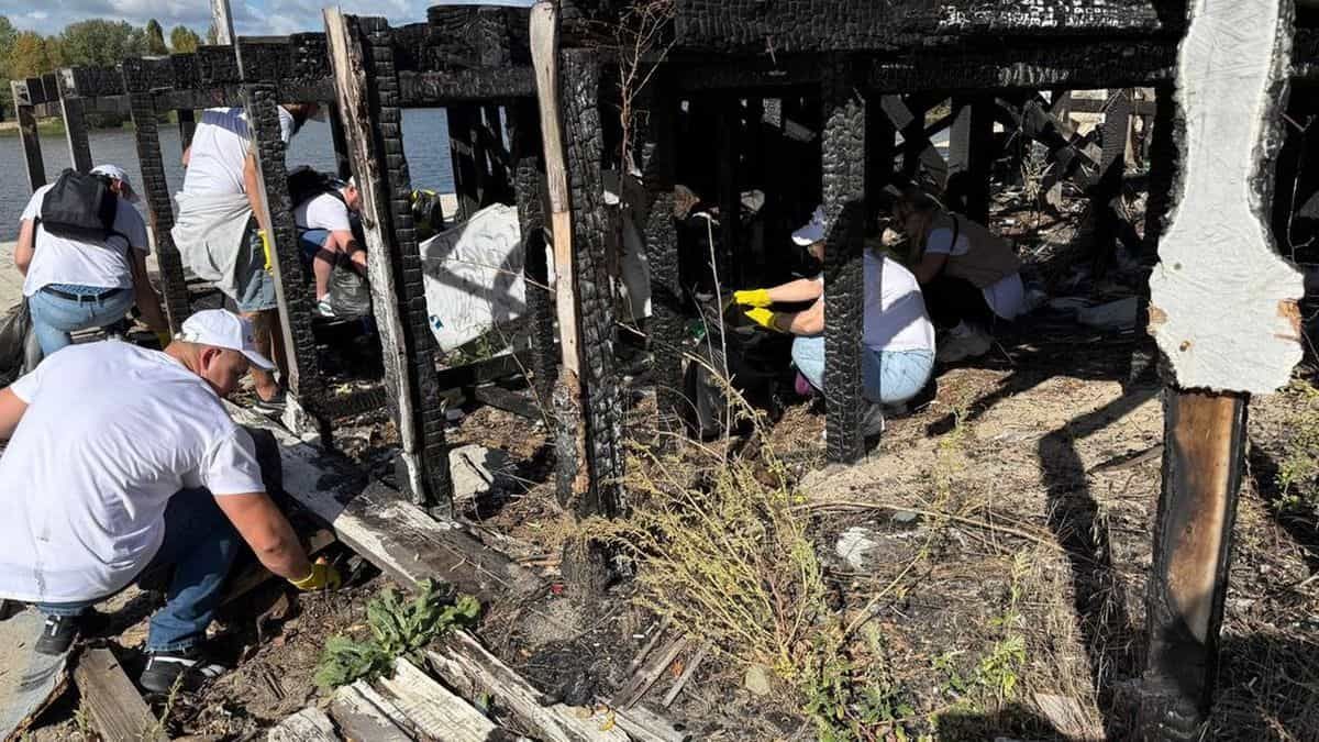 Люди в білих кепках збирають сміття серед обгорілих дерев’яних конструкцій біля води