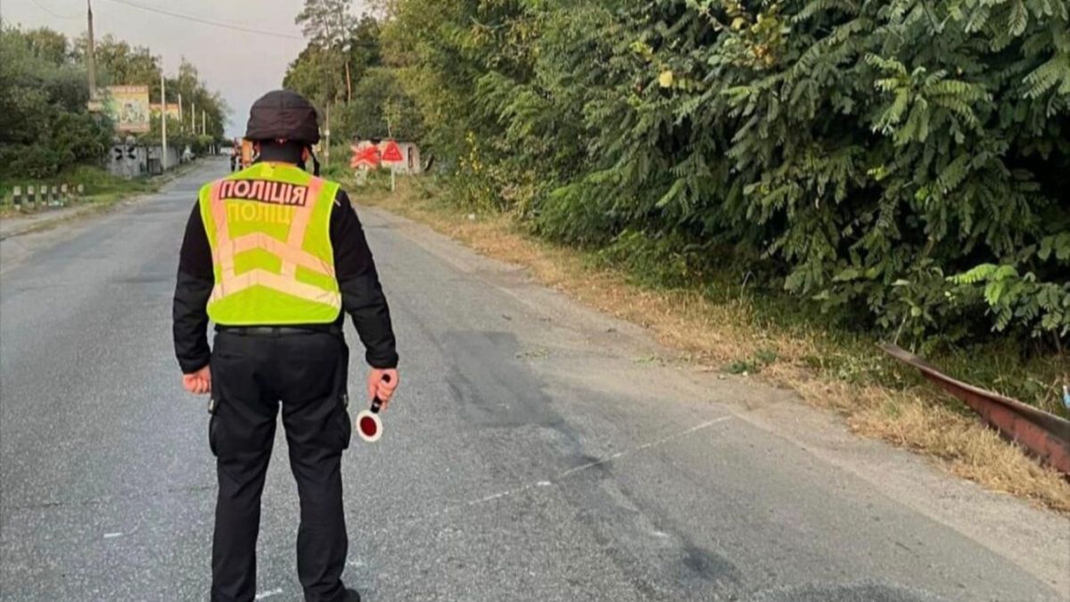 Поліцейський у чорній формі та жовтому світловідбивному жилеті стоїть на сільській дорозі біля лісосмуги