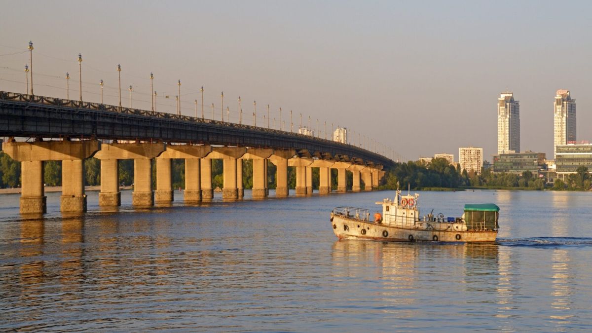 Міст Патона через Дніпро в Києві — вид збоку на бетонні опори мосту з баржею на річці та панорамою міста