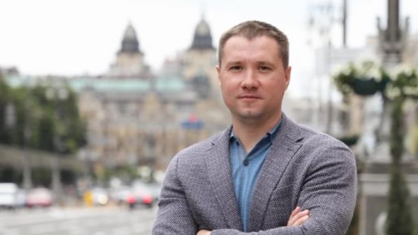 Чоловік у піджаку стоїть на тлі міського проспекту з розмитими будівлями та автомобілями