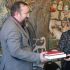 Чоловік із бородою і жінка передають одне одному дві червоні книги на тлі керамічного панно з князівськими воїнами та деревом