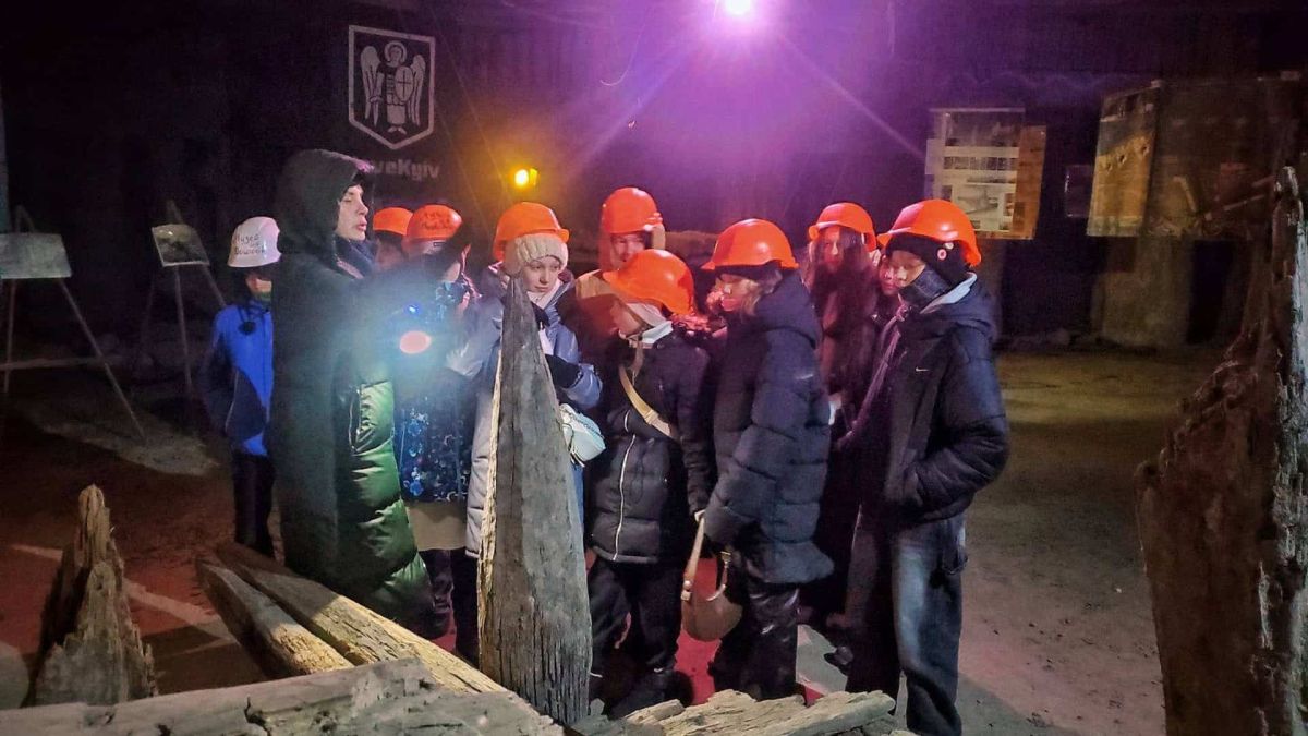 Діти в помаранчевих захисних касках та екскурсовод оглядають старовинні дерев'яні конструкції в підземній частині Музею на Поштовій площі у Києві